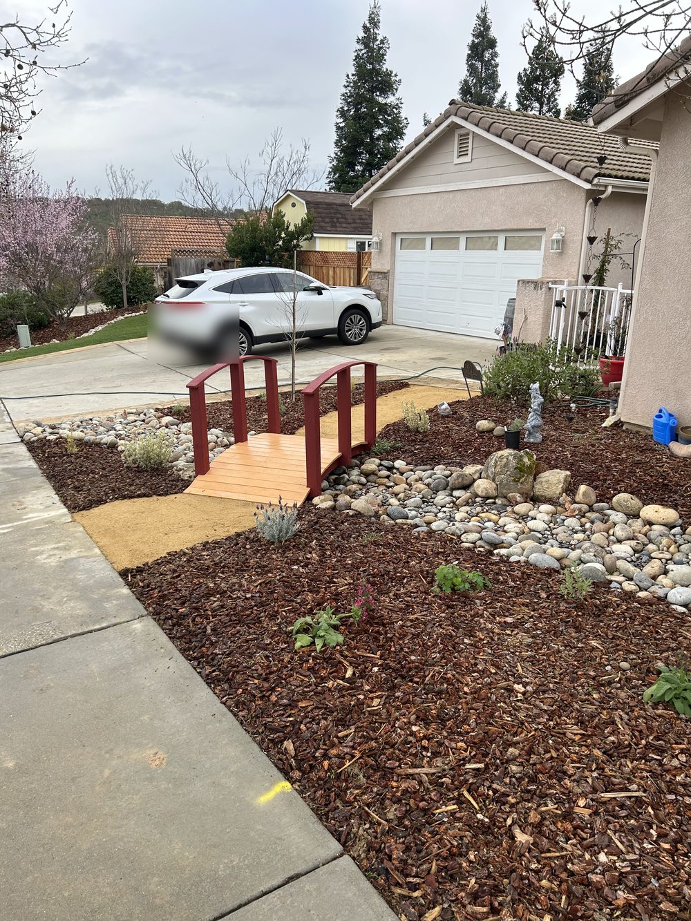 A small red wooden footbridge over a dry creek bed of river rock, with bark mulch and young plants around it.