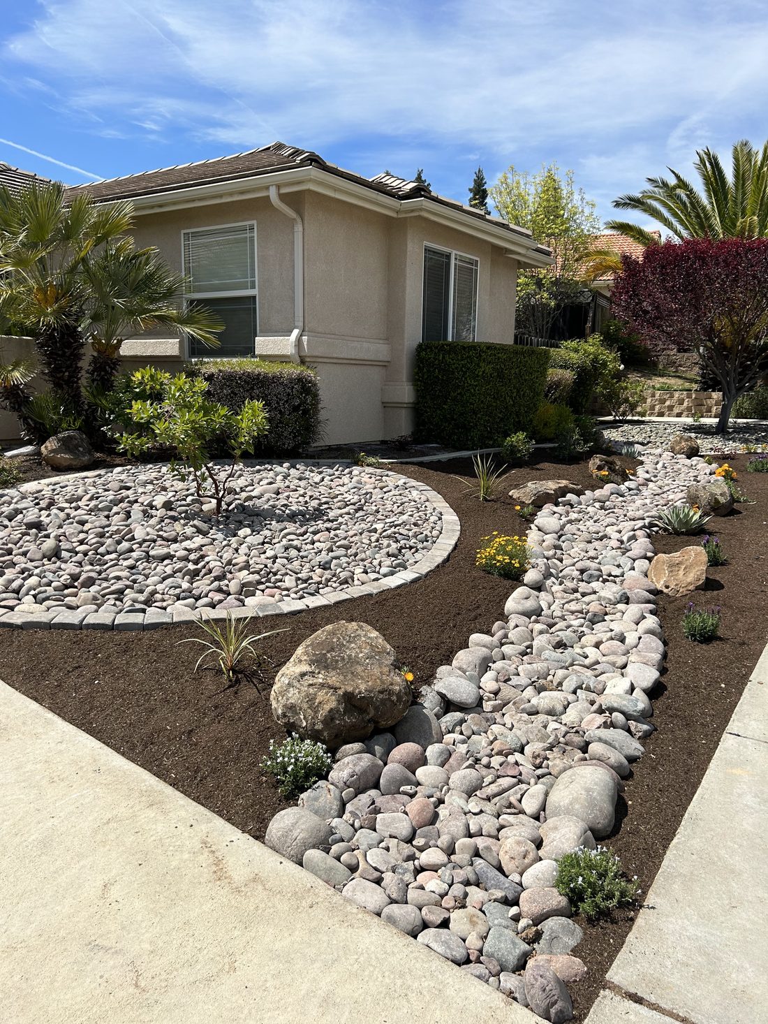 A front yard with a curving dry creek bed of river rock, a round rock bed edged in pavers, dark bark mulch, boulders and new plantings in front of a stucco house.
