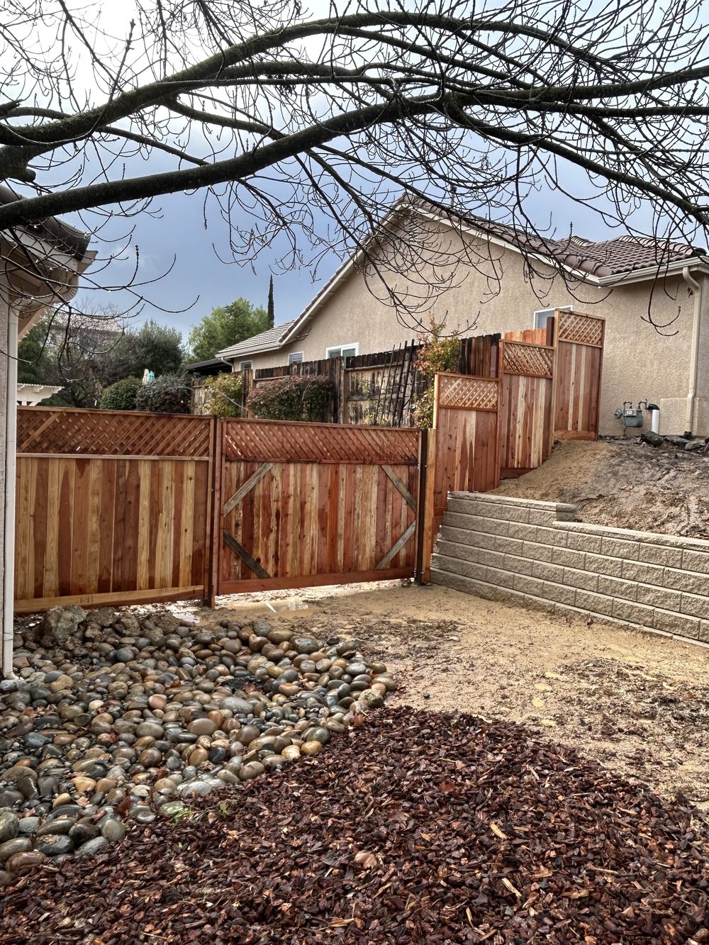 A new redwood fence and gate stepping down a slope beside a block retaining wall, with river rock and bark mulch in front.