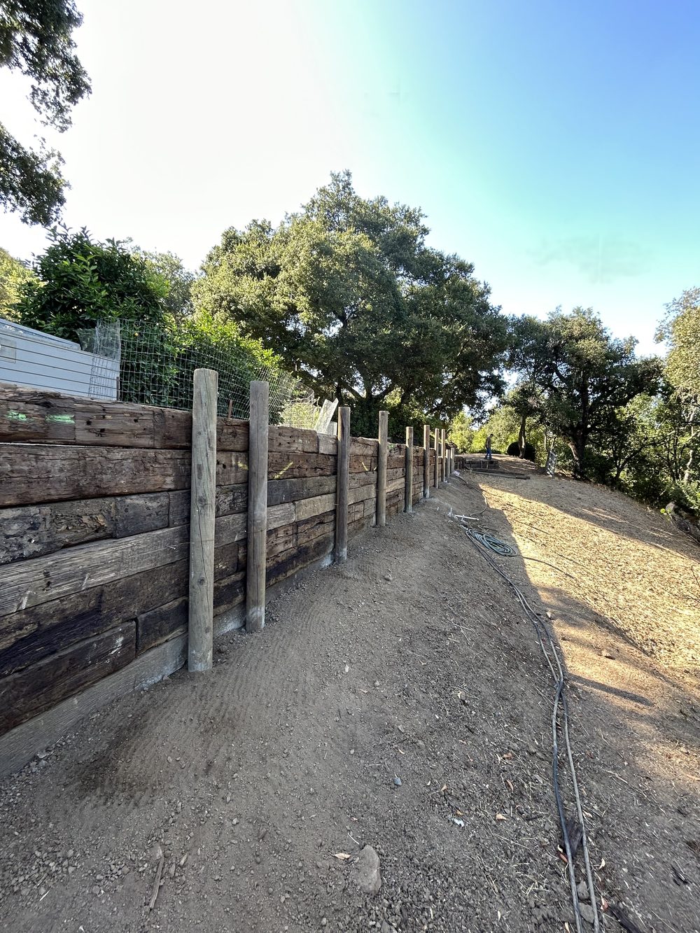 A long timber retaining wall with posts along a graded dirt slope under oak trees.